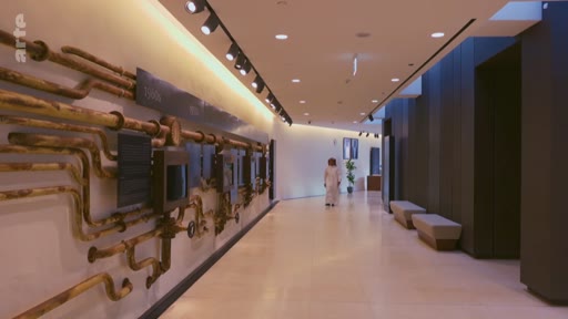 Une personne marche dans un long couloir à l'architecture contemporaine, orné d'une installation artistique murale composée de tuyaux en métal, évoquant un environnement industriel ou un musée technique.