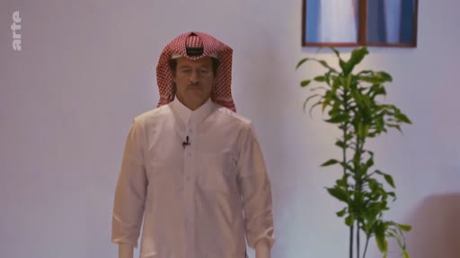 Un homme en tenue traditionnelle saoudienne, comprenant une chemise blanche et un keffieh rouge et blanc, est filmé sur un plateau de télévision avec une plante en arrière-plan. Il porte un micro-cravate, suggérant une interview ou une intervention médiatique.