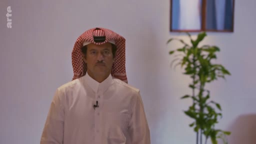 Un homme en tenue traditionnelle qatarie, incluant une ghutra rouge et blanche et un thobe blanc, est filmé en plan moyen dans un studio de télévision. Il porte un micro-cravate, suggérant une interview ou une intervention médiatique sur le Qatar.
