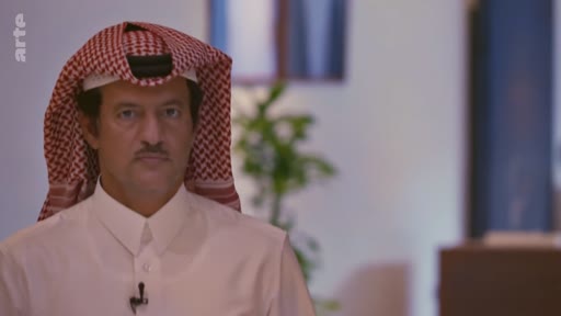 Un homme portant une ghutra et un thobe blanc est filmé en gros plan lors d'une interview télévisée. Il s'exprime face caméra dans un cadre formel, probablement pour un reportage sur l'Arabie saoudite.