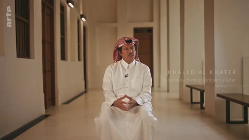 Khaled Al Khater, identifié par un bandeau, est interviewé dans un couloir à l'architecture traditionnelle au Qatar. Il porte une tenue traditionnelle qatarienne, incluant une thobe blanche et une ghutra.