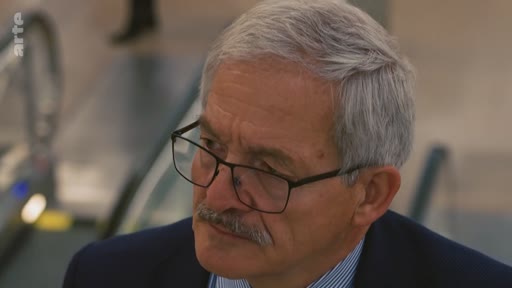 Gros plan sur un homme d'âge mûr aux cheveux gris et portant des lunettes, lors d'une interview télévisée.