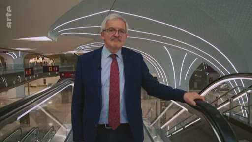 Un présentateur de télévision, portant une veste bleue et une cravate rouge, se tient sur un escalator dans une gare ou un aéroport à l'architecture moderne et futuriste. Le logo de la chaîne Arte est visible en haut à gauche.