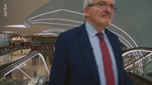 Un homme âgé, portant des lunettes, un costume bleu et une cravate rouge, est filmé en train de descendre un escalator dans une gare ou un aéroport à l'architecture moderne et lumineuse.