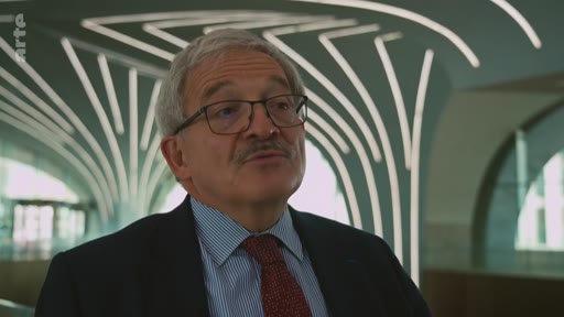 Un homme d'âge mûr, portant des lunettes, un costume sombre et une cravate, est interviewé dans un cadre architectural moderne avec des lignes lumineuses en arrière-plan.