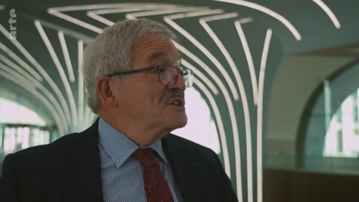 Un homme âgé, portant des lunettes, une chemise à rayures et une cravate, est interviewé dans un espace intérieur doté d'une architecture contemporaine avec des structures lumineuses en forme d'arbres.