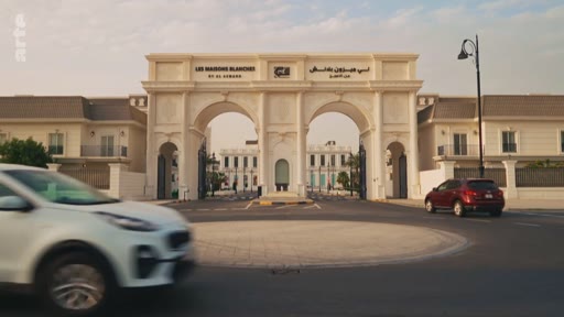 Vue extérieure de l'entrée monumentale du complexe résidentiel Les Maisons Blanches à Dubaï, avec des voitures circulant sur le rond-point au premier plan. Le site est identifié par une inscription bilingue en français et en arabe sur l'arche d'entrée.