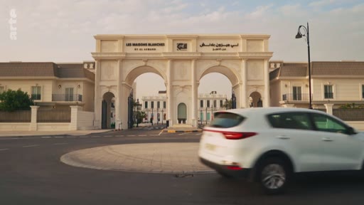 Une voiture circule devant l'entrée principale du complexe résidentiel Les Maisons Blanches, un projet immobilier situé aux Émirats arabes unis. L'architecture de la porte monumentale est visible avec des inscriptions en français et en arabe.
