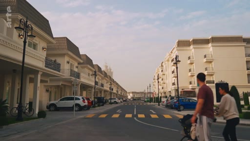 Un couple avec une poussette marche dans une rue bordée de maisons et d'immeubles résidentiels modernes, probablement dans un quartier récent ou une zone de développement urbain.