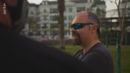 Un homme chauve portant des lunettes de soleil à verres bleus est filmé de profil lors d'une discussion en extérieur, avec un bâtiment en arrière-plan.