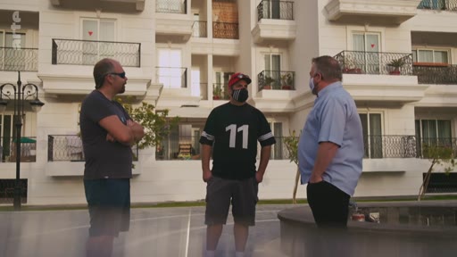 Trois hommes, dont l'un porte un maillot avec le numéro 11, discutent à l'extérieur devant un immeuble résidentiel moderne. La scène semble être une séquence de reportage ou d'interview en extérieur.