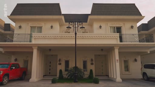 Vue de face d'un complexe résidentiel composé de deux maisons jumelées de style classique avec des balcons et des voitures garées devant. L'architecture présente des façades claires et des toits en pente, typiques des quartiers résidentiels modernes.