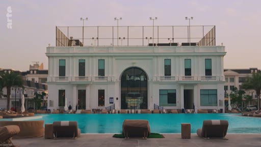 Vue d'ensemble d'un complexe hôtelier ou résidentiel haut de gamme avec une grande piscine turquoise et des chaises longues, surmonté d'un terrain de sport sur le toit.