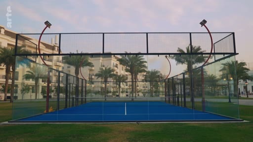 Vue d'ensemble d'un terrain de padel moderne avec une surface bleue et des parois en verre, situé dans un environnement extérieur avec des palmiers et des bâtiments en arrière-plan.