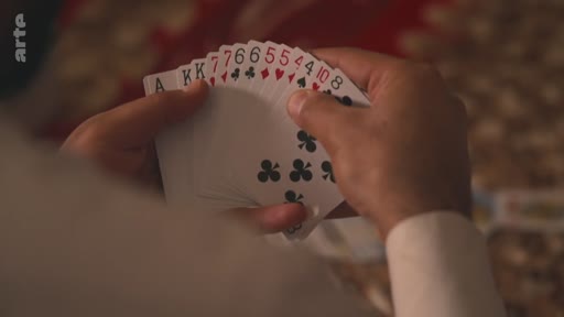 Gros plan sur les mains d'une personne tenant un éventail de cartes à jouer lors d'une partie. On distingue les valeurs des cartes, notamment des rois, des sept, des six, des cinq, des quatre, un dix et un huit.