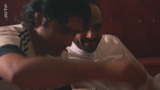 Deux hommes, dont l'un porte une tenue traditionnelle, sont en pleine conversation dans un intérieur sombre. L'ambiance est intime et feutrée.