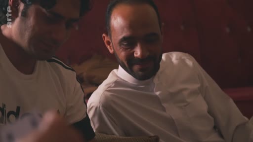 Deux hommes, dont l'un porte une tenue traditionnelle, sont assis côte à côte et discutent dans un cadre intérieur informel.