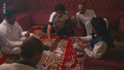 Un groupe d'hommes est assis en cercle sur un tapis rouge pour jouer aux cartes dans une pièce aux murs recouverts de banquettes. L'ambiance est décontractée, typique d'un moment de loisir entre amis.