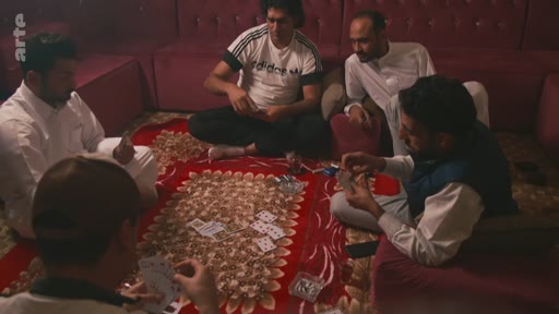 Un groupe d'hommes est assis en cercle sur un tapis rouge pour jouer aux cartes dans une pièce dotée de banquettes, dans une ambiance décontractée. La scène montre une activité de loisir en intérieur.