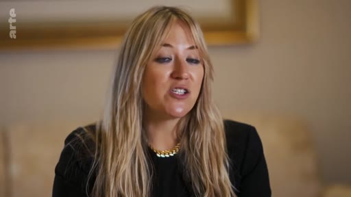 Une femme blonde aux cheveux longs, vêtue d'un haut noir et d'un collier doré, est filmée en gros plan lors d'une interview télévisée.