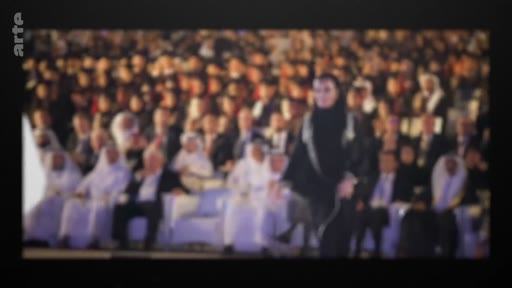 Une femme vêtue d'une abaya noire traverse une salle remplie de spectateurs lors d'une conférence ou d'une cérémonie officielle dans un pays du Golfe. L'assistance est composée majoritairement d'hommes en tenue traditionnelle, suggérant un événement politique ou diplomatique.