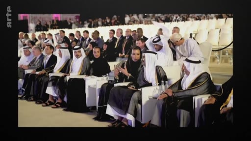 Un large public, incluant des hommes en thobe et ghutra et des femmes en abaya, est assis dans une salle lors d'une cérémonie ou d'un événement officiel au Moyen-Orient.