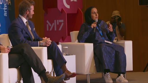 David Beckham participe à une discussion lors d'un événement au centre de design M7 à Doha, au Qatar. Il est assis aux côtés d'une femme portant un hijab, tous deux prenant la parole devant un public.