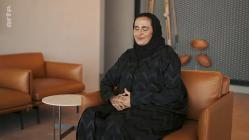 Une femme portant un hijab noir et une abaya est assise dans un fauteuil en cuir marron lors d'une interview télévisée. Elle a les yeux fermés et les mains jointes, dans un décor minimaliste et moderne.