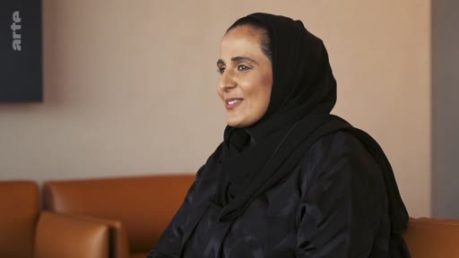 Une femme portant un hijab noir est filmée en plan moyen lors d'une interview télévisée. Elle est assise dans un intérieur sobre avec des fauteuils en cuir.