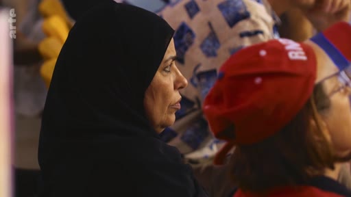 Une femme portant un hijab noir est filmée de profil lors d'un rassemblement public, aux côtés d'une personne portant une casquette rouge avec l'inscription RN.