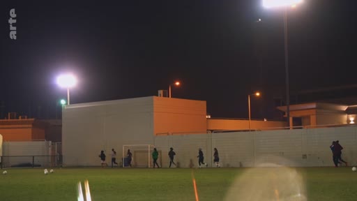 Un groupe de personnes effectue un entraînement physique sur un terrain de football en extérieur, sous l'éclairage artificiel des projecteurs, lors d'une séance nocturne.