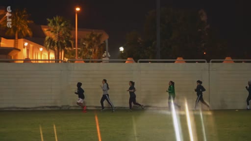 Un groupe de jeunes femmes court sur une piste ou un terrain de sport en extérieur, de nuit, sous l'éclairage des projecteurs. Cette scène illustre la pratique sportive féminine et l'entraînement physique en soirée.