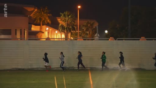 Un groupe de jeunes femmes court en file indienne sur un terrain de sport éclairé en extérieur, lors d'une séance d'entraînement nocturne.