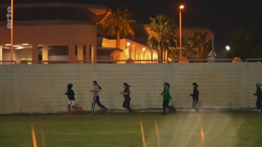 Un groupe de jeunes femmes court sur une piste d'athlétisme en extérieur lors d'une séance d'entraînement nocturne, avec un bâtiment éclairé en arrière-plan.