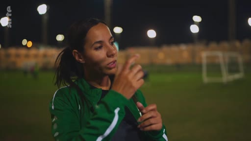 Une jeune femme en tenue de sport verte est interviewée sur un terrain de football éclairé en extérieur, probablement dans le cadre d'un reportage sur le sport féminin.