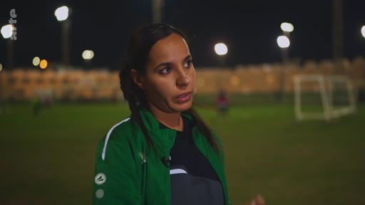 Une femme, portant une veste de sport verte, est interviewée sur un terrain de football éclairé en extérieur lors d'une séquence de reportage.