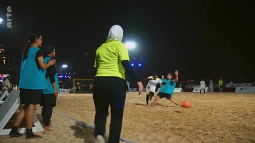 Des joueuses de football, dont certaines portent le hijab, participent à un match sur un terrain de sable en extérieur lors d'une soirée éclairée. L'image montre une action de jeu avec une joueuse en maillot bleu tentant de contrôler le ballon sous le regard d'une arbitre ou d'une entraîneuse en tenue jaune.