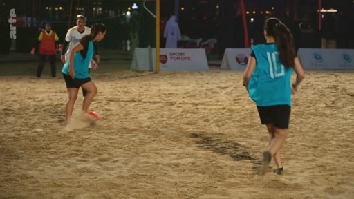 Des joueuses de football en maillots bleus s'entraînent sur un terrain de sable lors d'une session sportive en extérieur. On peut apercevoir une banderole avec l'inscription 'SPORT FOR LIFE' en arrière-plan.
