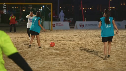 Des joueuses de football en maillots bleus disputent un match sur un terrain de sable en extérieur lors d'un événement nocturne intitulé 'Sport for Life'.