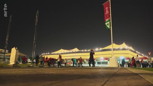 Vue extérieure nocturne du stade Al-Bayt à Al Khor, au Qatar, lors de la Coupe du Monde de la FIFA 2022, avec des spectateurs marchant sur le parvis sous les drapeaux officiels de l'événement.