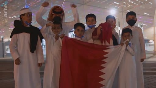 Un groupe d'enfants et un homme en tenue traditionnelle qatarienne posent ensemble en tenant le drapeau du Qatar lors d'un événement en extérieur. La scène illustre la culture et le patriotisme au Qatar.