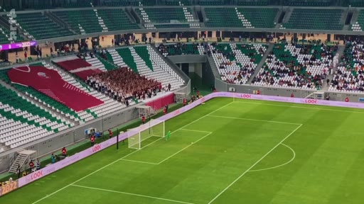 Vue en plan large d'un stade de football lors d'un match, mettant en évidence un tifo déployé dans les tribunes représentant un maillot de sport. L'ambiance est celle d'une rencontre sportive professionnelle avec des supporters présents dans les gradins.
