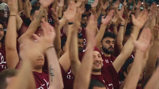 Une foule de supporters portant des t-shirts bordeaux avec l'inscription 'QATAR' lèvent les bras en l'air dans un stade, illustrant l'ambiance lors d'un événement sportif au Qatar.