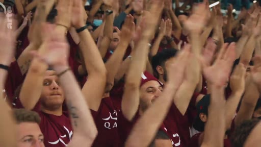 Une foule de supporters portant des t-shirts bordeaux avec le logo 'QA' applaudit en levant les bras lors d'un événement sportif, probablement au Qatar.