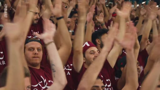 Une foule de supporters portant des t-shirts bordeaux avec l'inscription 'QATAR' acclame lors d'un événement sportif, probablement une rencontre de football au Qatar.