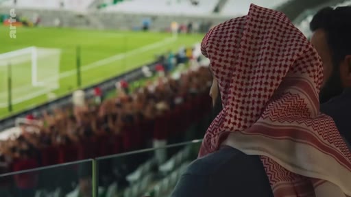 Un spectateur portant un keffieh traditionnel observe un match de football dans un stade depuis une loge VIP. En arrière-plan, on aperçoit la foule des supporters et le terrain de jeu.