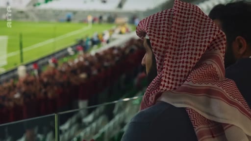 Le prince héritier d'Arabie saoudite, Mohammed ben Salmane, est filmé de profil alors qu'il assiste à un match de football dans un stade. Il porte une ghutra traditionnelle rouge et blanche.
