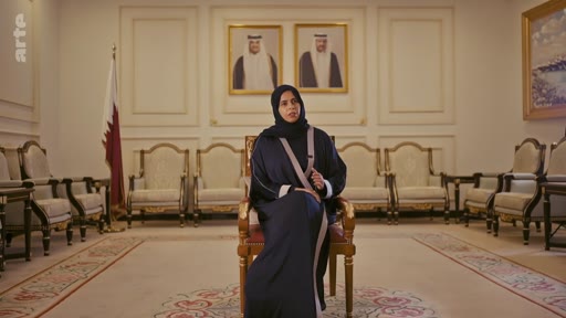 Une femme portant une abaya et un hijab est assise dans un salon officiel au Qatar, avec le drapeau du Qatar et des portraits de dirigeants en arrière-plan. Cette interview se déroule dans un cadre formel, probablement dans un bâtiment gouvernemental ou diplomatique.