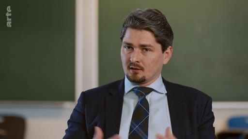 Un homme en costume et cravate s'exprime face caméra lors d'une interview télévisée, probablement dans un cadre académique ou professionnel.