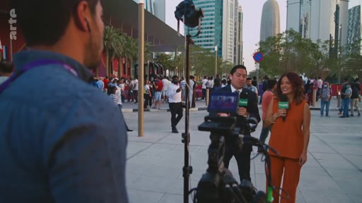 Deux journalistes, un homme et une femme, présentent un reportage en direct devant des gratte-ciels modernes, probablement à Doha au Qatar, avec une foule en arrière-plan.